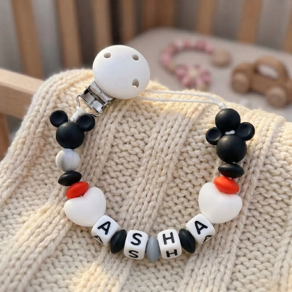 A beaded pacifier clip or baby accessory with a white silicone top, black Mickey-shaped beads, white letter beads spelling A S H A, and small red and gray accents, displayed on a cream knit blanket.