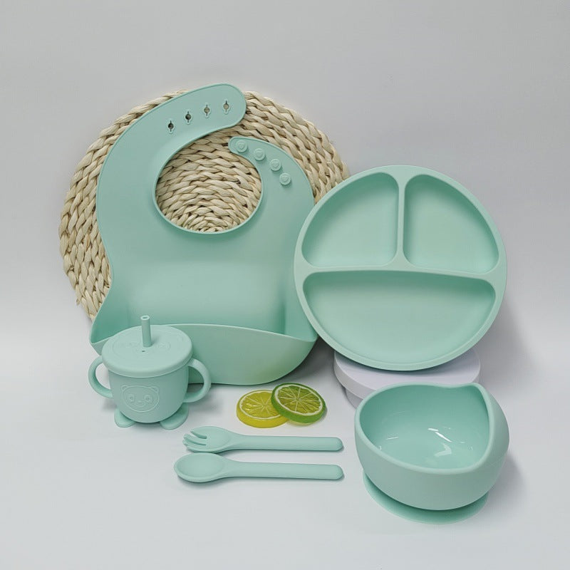 Mint green children's dining set including a bib, plate, cup, bowl, and utensils on a light gray background.