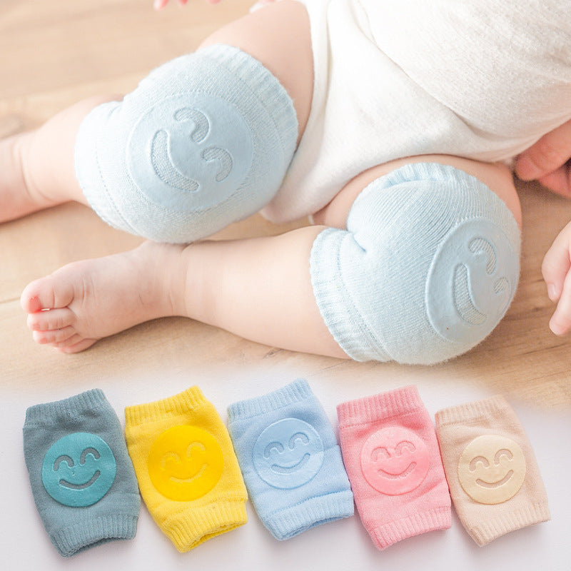 Baby wearing light blue knee pads with smiley face design, surrounded by additional knee pads in different colors on a light background.