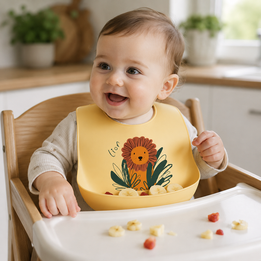 A smiling baby sitting in a wooden high chair wearing a yellow silicone bib with a colorful flower illustration and the word 'lion'.