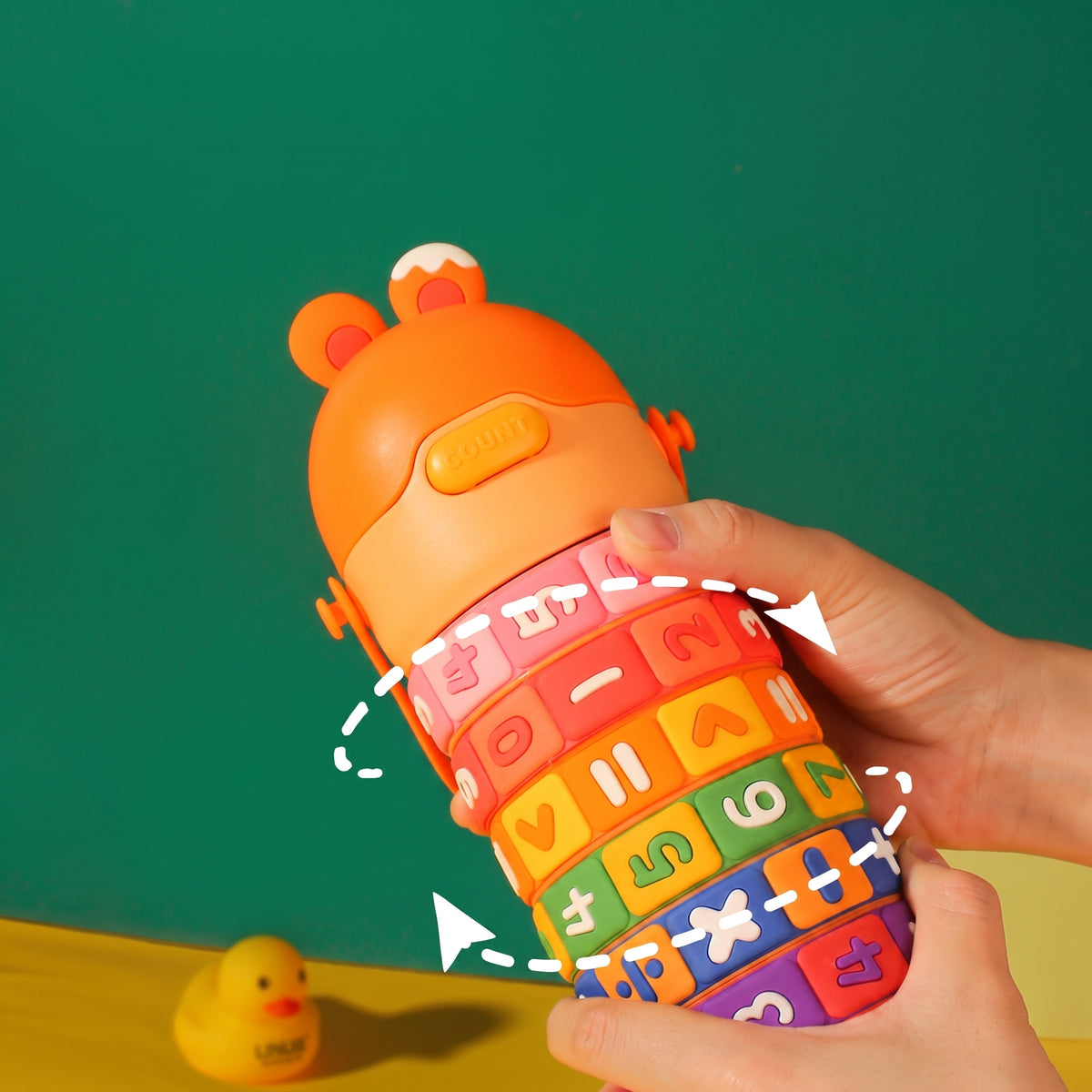 A child's math learning travel cup held by two hands against a green background. The cup features a rotating orange bear-shaped top and colorful rings with mathematical symbols and numbers.