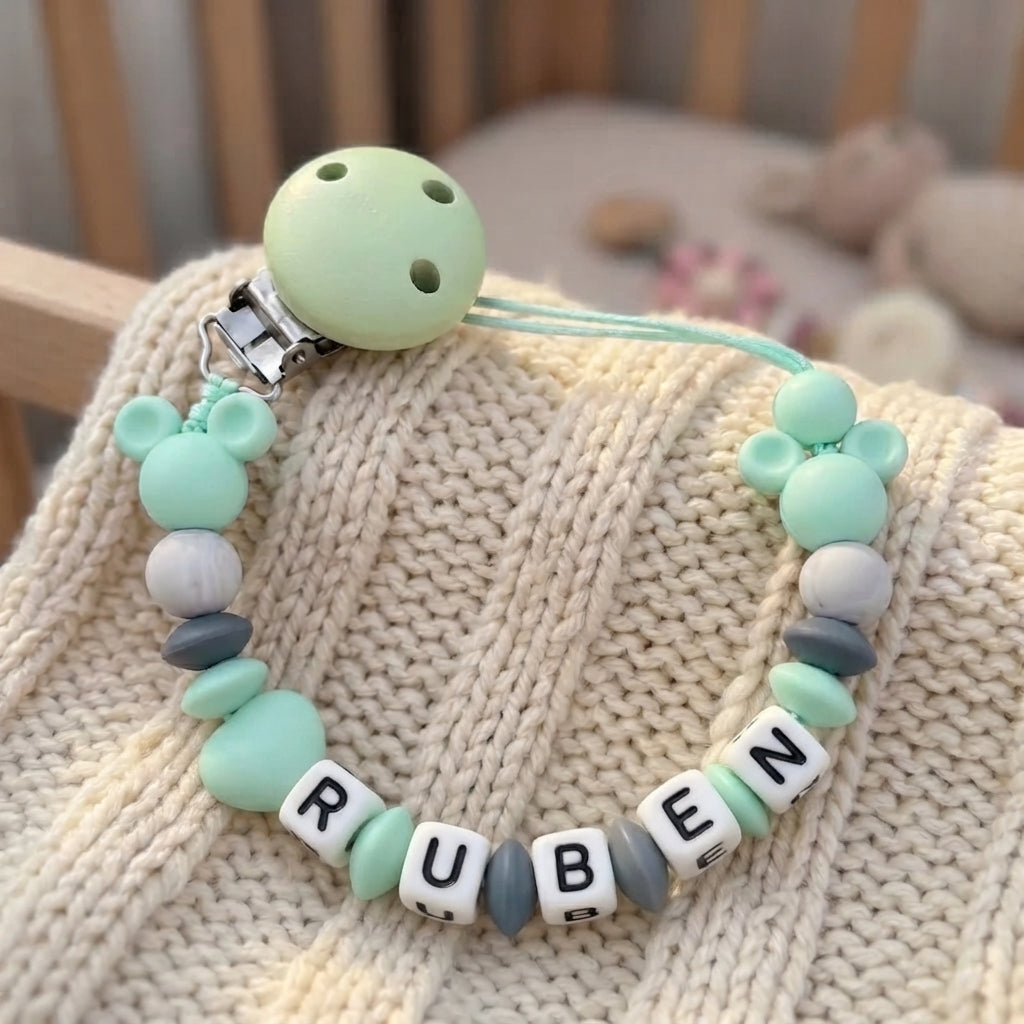 A personalized baby pacifier clip with mint green, white, and gray silicone beads spelling 'RUBEN' on white letter cubes, resting on a cream-colored knit blanket.