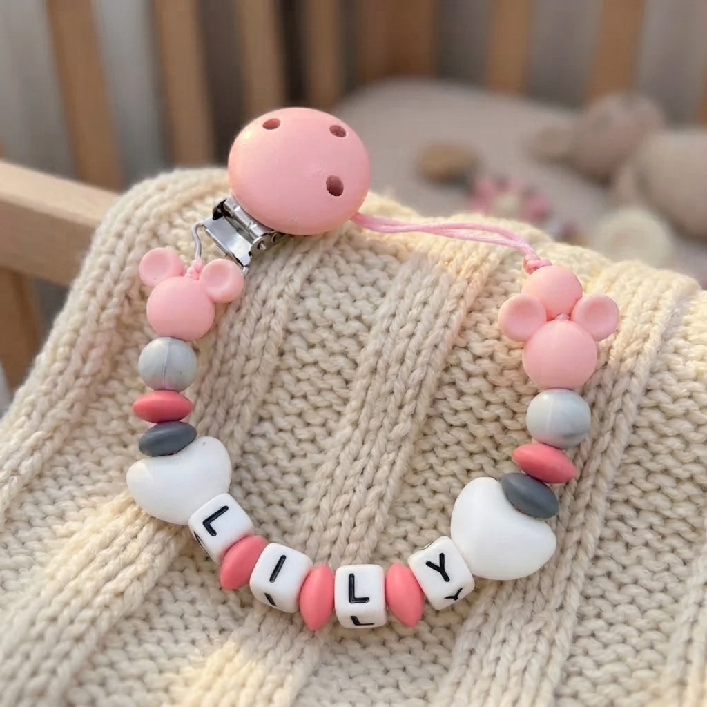 A personalized baby pacifier clip with pink silicone Mickey Mouse ears, white and pink beads, and name beads spelling 'LILY' on a beige knit blanket.