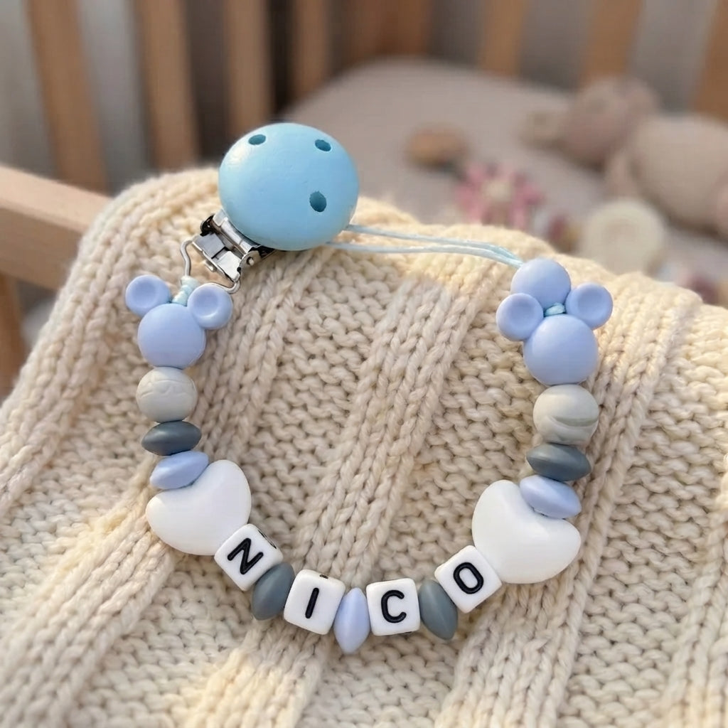 A baby pacifier clip with a light blue silicone ball, a silver metal clip, and a beaded chain featuring blue and white silicone beads, marble accents, and letter tiles spelling 'NICO', resting on a cream-colored knit blanket.
