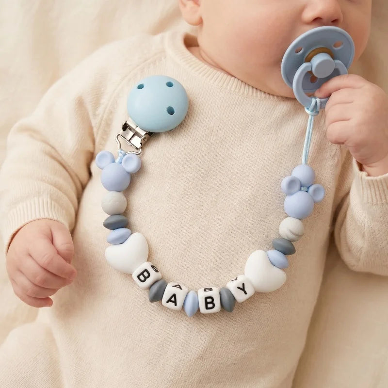 Baby holding a pacifier with a colorful teething ring attached, wearing a beige outfit.