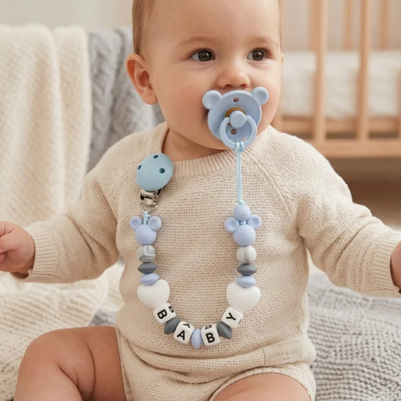 Baby wearing a blue pacifier with a teething chain in a cozy room.