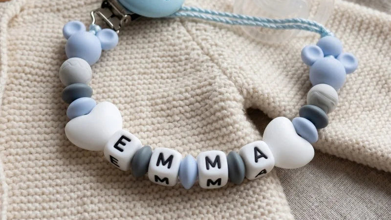 A silicone baby teether bracelet with letter beads spelling 'EMMA' and heart-shaped beads, resting on a textured beige knit blanket.