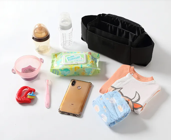 A baby packing kit arranged on a white background, featuring a black diaper bag organizer, a gold smartphone, a blue patterned diaper, a white and peach baby bib, a pack of wet wipes, a pink bowl, a pink toothbrush, a red teething toy, and two baby bottles.