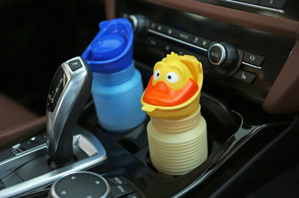 A yellow duck-shaped car cup holder holder with an orange beak and white eyes, holding a beige ribbed bottle, shown next to a blue duck-shaped cup holder in a car's center console.