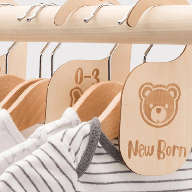 Close-up of a wooden baby clothes hanger rack with engraved labels including 'New Born' and '0-3'. A white and gray striped baby shirt is hanging on the front wooden hanger, which has a teddy bear face engraving.