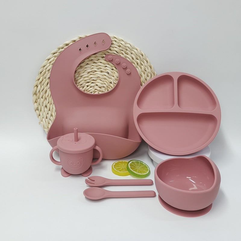 Pink children's dining set including a bib, plate, cup, bowl, and utensils on a white background.