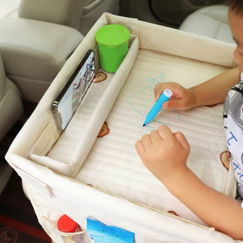 A child sitting in a car uses a padded cream-colored car activity table. The table has a built-in tray holding a smartphone and a green cup, with a child drawing on the padded surface using a blue marker.