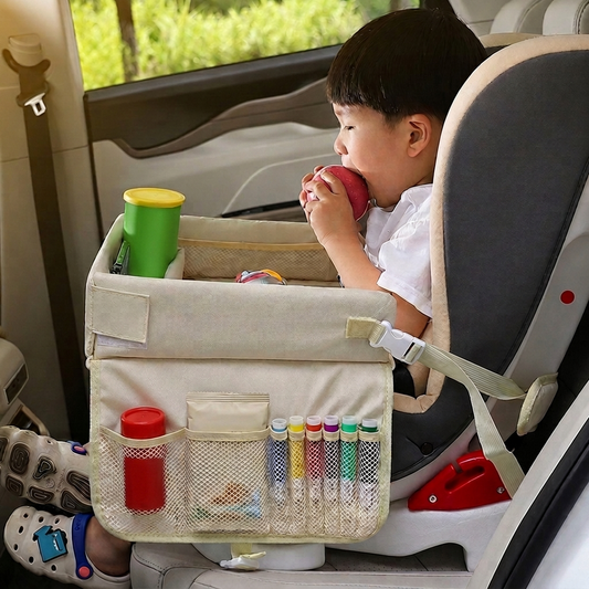A child sitting in a car seat uses a beige fabric car-side organizer bag mounted on the seat, holding a red apple and drinking from a green cup. The bag has mesh pockets holding pens and small items.